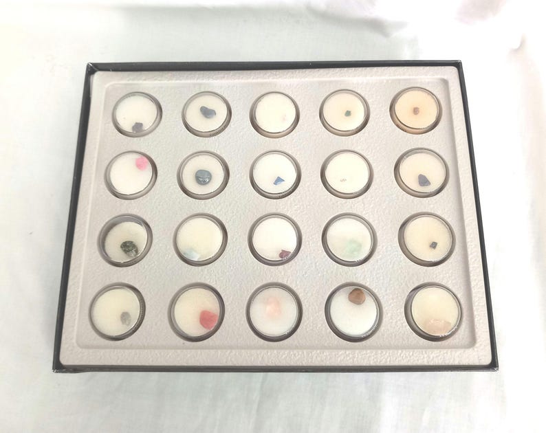 Puede incluir: Una bandeja rectangular negra con 20 peque&ntilde;os recipientes redondos y transparentes. Cada recipiente contiene una peque&ntilde;a gema de colores colocada en cera blanca. Las gemas son de varios colores, incluyendo rosa, azul, verde y marr&oacute;n.