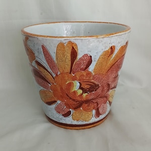 Op de afbeelding: Een taps toelopende keramische plantenpot met een witte basis met een gespikkelde textuur en een groot, handgeschilderd bloemmotief in tinten oranje, roest en geel. De rand en de voet zijn geaccentueerd met een oranjebruine band, binnenkant is wit.