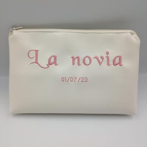 Puede incluir: Bolsa blanca con cremallera con el texto "La novia" y la fecha "01/07/22" bordada en rosa.