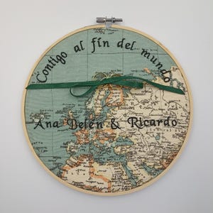 May include: A wooden embroidery hoop featuring a vintage world map design. The map is in shades of blue, green, and brown. The text "Contigo al fin del mundo" is embroidered in black thread around the edge of the map. The names "Ana Belen & Ricardo" are embroidered in black thread below the map. A green ribbon is tied around the hoop, with two gold rings on top of the ribbon.