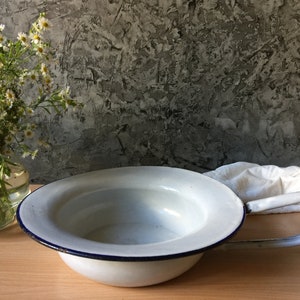 May include: White enamel bowl with a blue rim and a single handle. The bowl is sitting on a wooden surface.