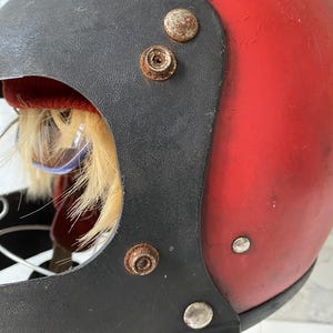 May include: A vintage-style red motorcycle helmet featuring a black visor trim with visible rust on the metal snap fasteners. The interior reveals a red fabric lining, a tuft of light-colored faux fur, and a glimpse of a red and white patterned fabric.