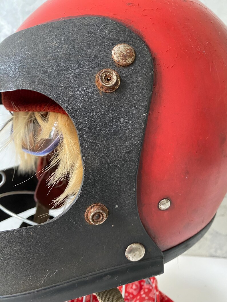 May include: A vintage-style red motorcycle helmet featuring a black visor trim with visible rust on the metal snap fasteners. The interior reveals a red fabric lining, a tuft of light-colored faux fur, and a glimpse of a red and white patterned fabric.