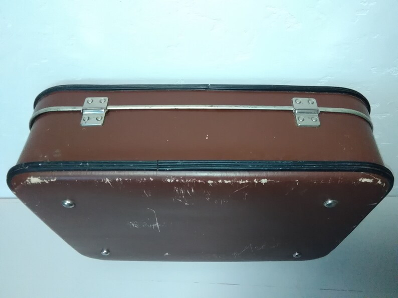 Może przedstawiać: Brązowa vintage walizka ze srebrnymi metalowymi zawiasami i czarną obw&oacute;dką. Walizka nosi ślady zużycia.
