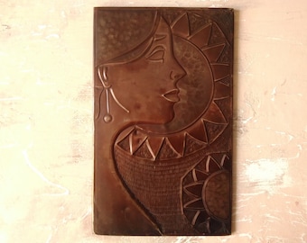 May include: A brown metal wall sculpture with a stylized image of a woman's face and a sun. The sculpture has a textured surface and a geometric design.