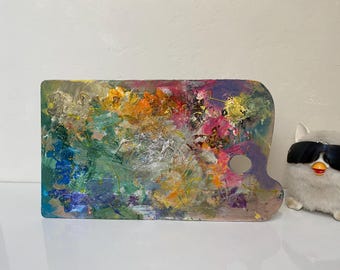 May include: A rectangular wooden artist's palette covered in a vibrant mix of paint. The palette features a thumb hole and is splattered with hues of blue, green, orange, pink, and yellow. The palette is on a white surface.