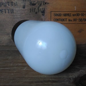 May include: A white, rounded, opaque glass bottle with a dark brown cap. The bottle is cylindrical with a rounded base and is set against a wooden background. The bottle is likely a container for liquids.