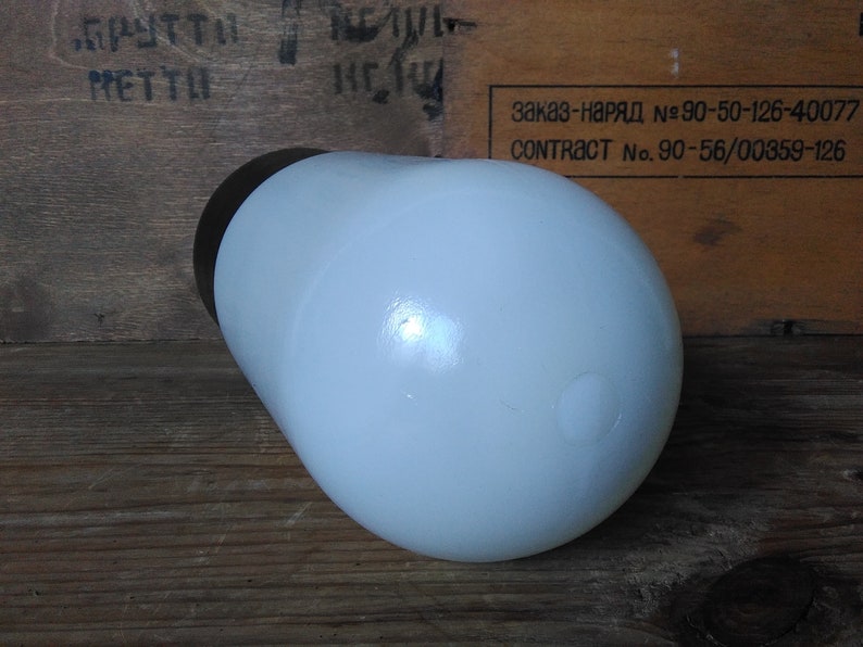 May include: A white, rounded, opaque glass bottle with a dark brown cap. The bottle is cylindrical with a rounded base and is set against a wooden background. The bottle is likely a container for liquids.