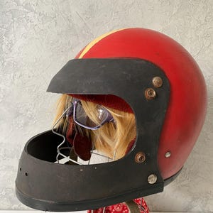 May include: A vintage red motorcycle helmet featuring a black visor and chin guard, accented by a yellow center stripe. The helmet is displayed on a wire stand with a red and white patterned bandana underneath and includes a pair of purple glasses.