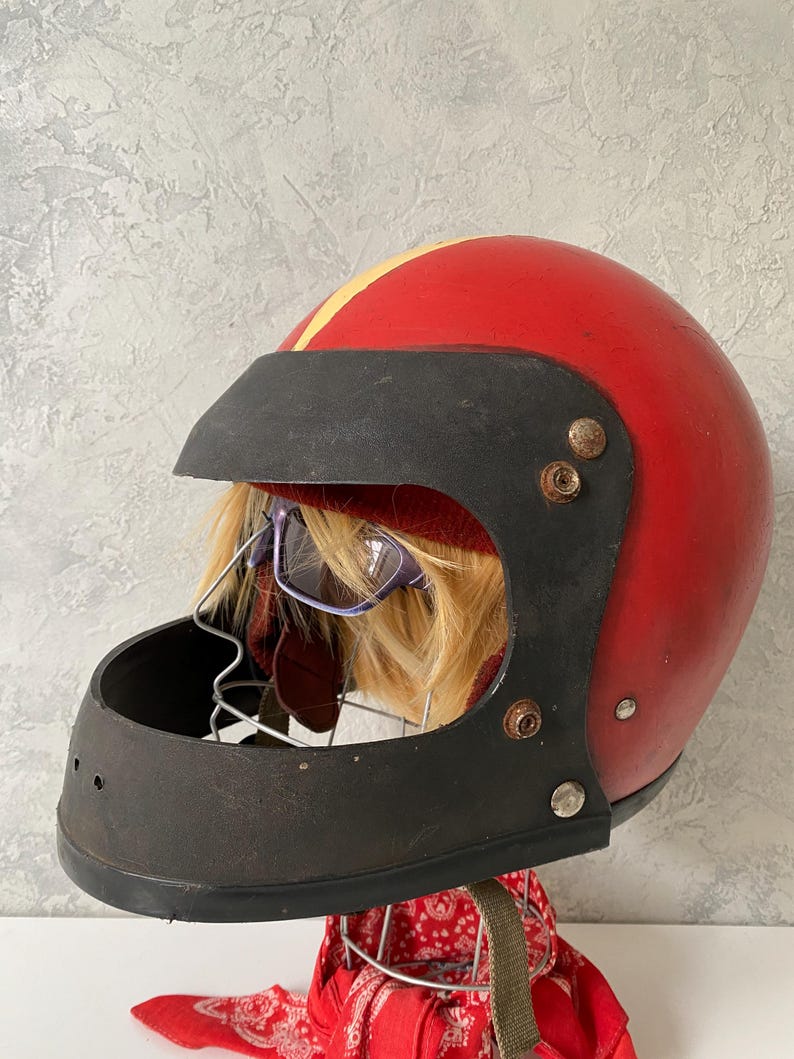 May include: A vintage red motorcycle helmet featuring a black visor and chin guard, accented by a yellow center stripe. The helmet is displayed on a wire stand with a red and white patterned bandana underneath and includes a pair of purple glasses.