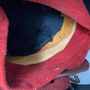 May include: The interior of a helmet featuring a bright red felt lining, a black fabric crown insert, and a tan leather sweatband. The structure includes a dark outer shell and a metallic buckle attachment visible at the lower edge of the assembly.