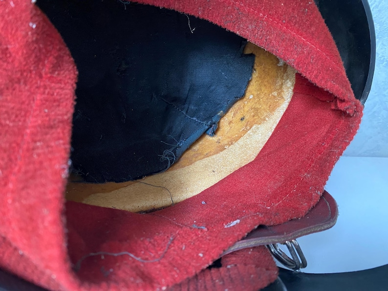 May include: The interior of a helmet featuring a bright red felt lining, a black fabric crown insert, and a tan leather sweatband. The structure includes a dark outer shell and a metallic buckle attachment visible at the lower edge of the assembly.