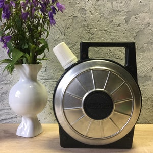 Könnte beinhalten: Eine vintage Thermosflasche in Schwarz und Silber mit einem weißen Deckel und einem schwarzen Griff. Die Thermosflasche hat ein kreisförmiges Design mit einer erhabenen Mitte und dem Wort "Thermos" darauf gedruckt.