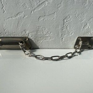 Può includere: Una catena di sicurezza per porta color argento, composta da due piastre di montaggio rettangolari e una catena. La serratura è installata su una superficie bianca, con un muro bianco strutturato sullo sfondo. La catena è composta da piccole maglie metalliche.