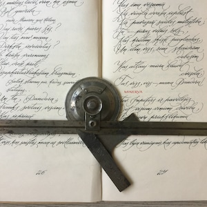 Puede incluir: Un libro abierto con texto escrito a mano en tinta negra sobre papel blanco. Un transportador de metal y una regla de metal se colocan sobre el libro abierto. El transportador tiene la palabra "MINERVA" impresa en rojo.