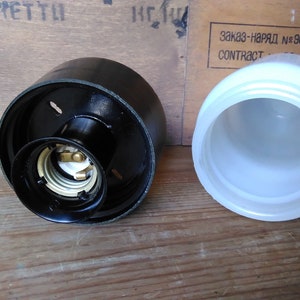 May include: A black lamp socket with a white interior and a separate white lampshade. The socket has two small holes and a brass-colored interior. The lampshade is cylindrical.