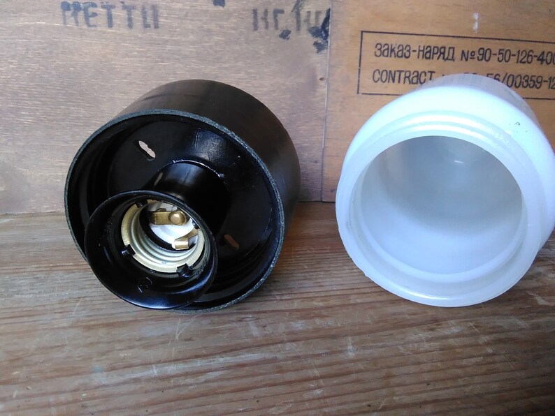 May include: A black lamp socket with a white interior and a separate white lampshade. The socket has two small holes and a brass-colored interior. The lampshade is cylindrical.