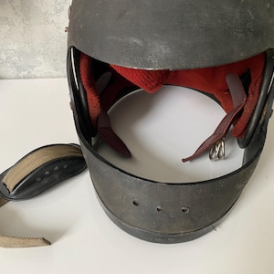 May include: A dark gray protective helmet with a wide front opening, featuring a red interior lining and a separate tan fabric chin strap with a black padded section. The helmet has visible metal rivets along the sides and ventilation holes.