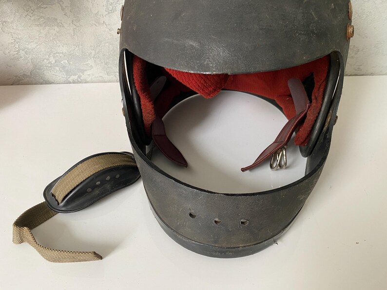 May include: A dark gray protective helmet with a wide front opening, featuring a red interior lining and a separate tan fabric chin strap with a black padded section. The helmet has visible metal rivets along the sides and ventilation holes.