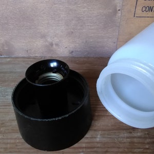 May include: A black lamp socket and a white glass lampshade are shown against a wooden background. The lamp socket is cylindrical with a black finish and a brass-colored interior. The lampshade is a jar shape.