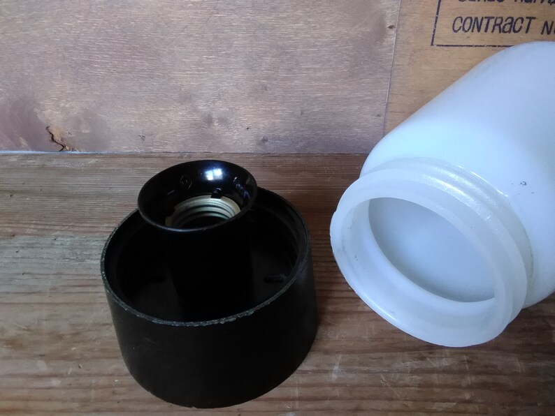 May include: A black lamp socket and a white glass lampshade are shown against a wooden background. The lamp socket is cylindrical with a black finish and a brass-colored interior. The lampshade is a jar shape.