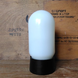 May include: A vintage white glass lamp with a rounded top and a black base. The lamp has a cylindrical shape and is set against a wooden background with text in Cyrillic script.