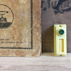 May include: A vintage, cream-colored doorbell with a green button and a rectangular nameplate. The doorbell is mounted on a wooden surface. The text "RIGA 1924" is visible on the surface.