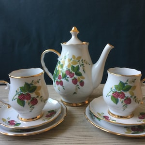 Könnte beinhalten: Ein weißes Porzellan-Tee-Set mit Himbeer-Design. Das Set beinhaltet eine Teekanne, zwei Tassen und zwei Untertassen. Die Tassen und Untertassen haben einen goldenen Rand.