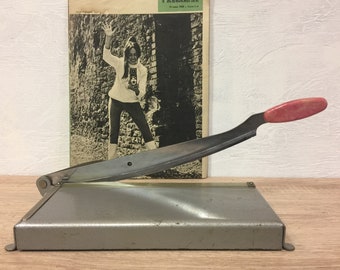 May include: A vintage paper cutter with a silver metal base and blade, and a red handle. The cutter is positioned in front of a black and white magazine cover featuring a woman holding a camera.