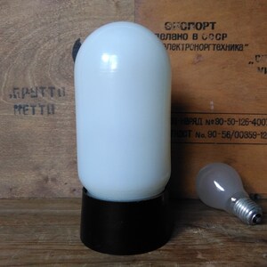May include: A vintage white glass lamp with a black base and a separate light bulb. The lamp has a cylindrical shape and sits on a wooden surface. The background features text in a foreign language.