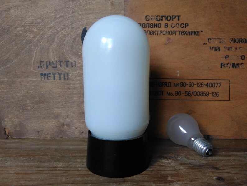 May include: A vintage white glass lamp with a black base and a separate light bulb. The lamp has a cylindrical shape and sits on a wooden surface. The background features text in a foreign language.