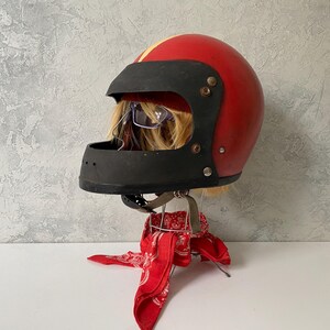May include: A red motorcycle helmet with a black face shield and a yellow stripe down the center. The helmet is displayed on a stand draped with a red and white patterned bandana, with blonde hair and glasses visible inside the helmet opening.