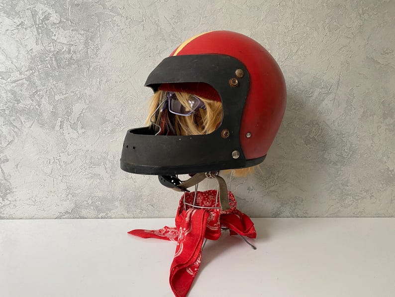 May include: A red motorcycle helmet with a black face shield and a yellow stripe down the center. The helmet is displayed on a stand draped with a red and white patterned bandana, with blonde hair and glasses visible inside the helmet opening.