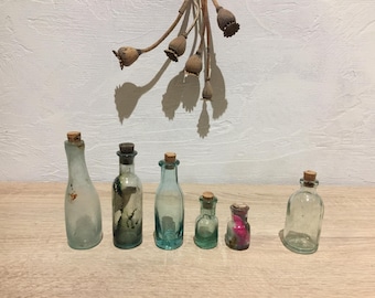 Puede incluir: Siete pequeñas botellas de vidrio con tapones de corcho. Las botellas son transparentes y tienen un tono azul claro. Están dispuestas en fila sobre una superficie de madera.