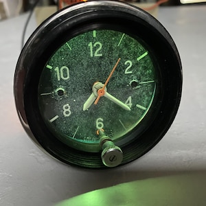 Puede incluir: Un reloj vintage con borde negro y esfera teñida de verde. La esfera del reloj muestra números y manecillas blancas, con un segundero naranja. Se ve un pequeño botón en la parte inferior. El reloj es probablemente un reloj de automóvil o de salpicadero.