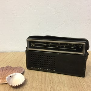 Könnte beinhalten: Ein schwarzes Vintage-Transistorradio mit einem silbernen Zifferblatt und einem Lederetui. Das Radio hat ein Lautsprechergitter an der Vorderseite und einen Drehknopf an der Seite.