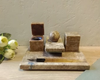 Op de afbeelding: Een marmeren bureauset met een pennenhouder, een inktpot en een klein doosje. De set is gemaakt van lichtbruin marmer met donkerbruine aderen. De pennenhouder bevat een zwarte pen met een gouden punt. De inktpot heeft een open deksel. Het kleine doosje heeft een gesloten deksel. De set staat op een lichtbruin houten oppervlak.