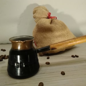 Könnte beinhalten: Eine schwarze und kupferne türkische Kaffeekanne mit einem Holzgriff, die auf einer Holzoberfläche steht, auf der Kaffeebohnen verstreut sind. Im Hintergrund befindet sich ein Jutesack mit einem roten Band.