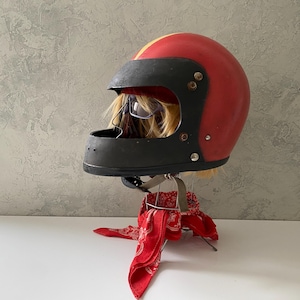 May include: A red motorcycle helmet with a yellow center stripe and a black face guard. The helmet features a matte black visor area with visible metal rivets and a chin bar with small ventilation holes, resting on a red and white patterned bandana.