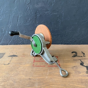 Peut inclure: Une meule à manivelle ancienne avec un couvercle de roue vert et une meule de couleur rouille. Le cadre métallique est argenté et la poignée est noire. L'outil est fixé sur une surface en bois.