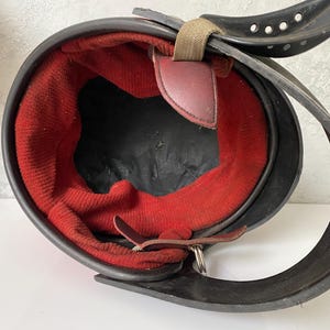 May include: A black protective helmet featuring a vibrant red fabric interior lining. The helmet includes a black chin strap with multiple circular ventilation holes, a tan fabric adjustment strap, and dark brown leather-like attachment tabs.