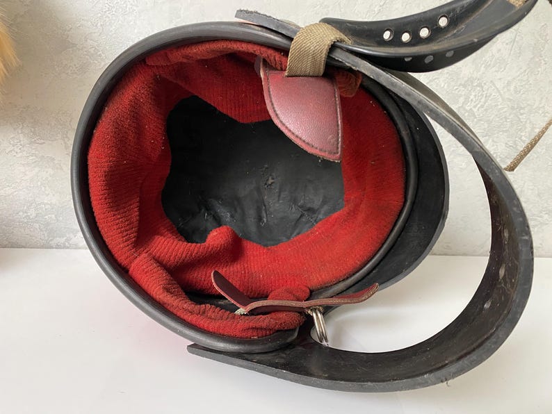 May include: A black protective helmet featuring a vibrant red fabric interior lining. The helmet includes a black chin strap with multiple circular ventilation holes, a tan fabric adjustment strap, and dark brown leather-like attachment tabs.