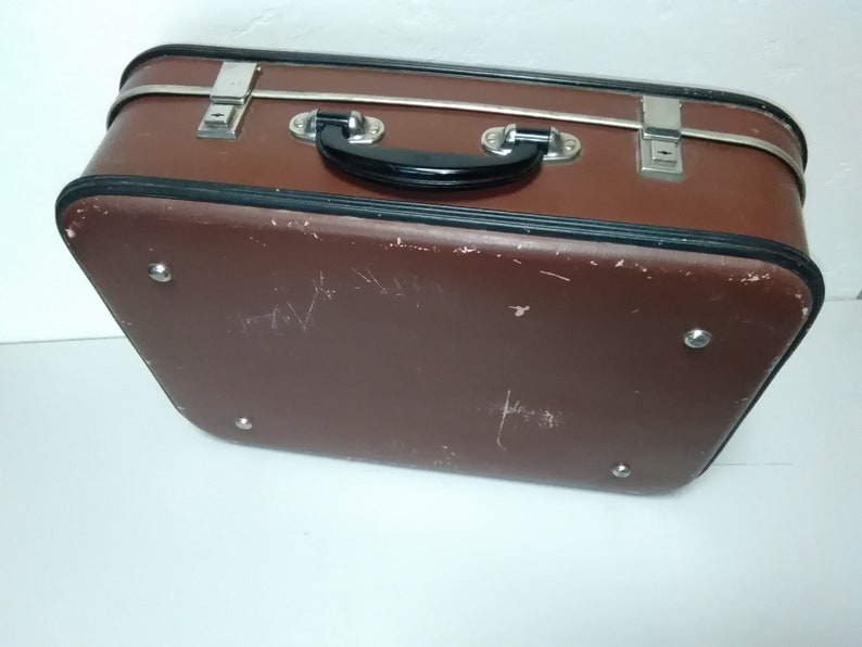 Może przedstawiać: Brązowa vintage'owa walizka z czarnym uchwytem i srebrnymi akcentami. Walizka ma zużyty wygląd z kilkoma zadrapaniami i otarciami.
