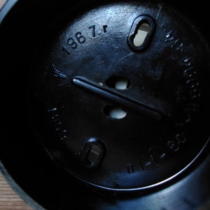 May include: A close-up shot of a black plastic object with a circular interior. The object has several holes and a raised central bar. The text "1967" and other markings are visible on the surface.