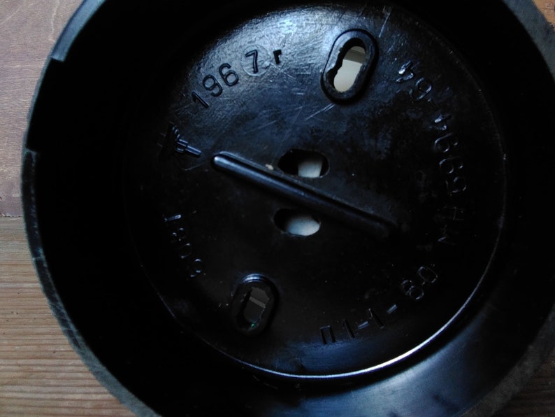 May include: A close-up shot of a black plastic object with a circular interior. The object has several holes and a raised central bar. The text "1967" and other markings are visible on the surface.