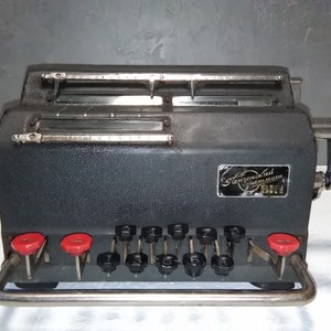 Op de afbeelding: Een zwarte vintage rekenmachine met rode knoppen en een zilveren handvat. De machine heeft een label met de tekst "Советский Счетмаш БКМ".