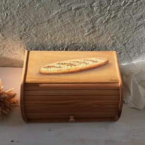 Könnte beinhalten: Eine Brotbox aus Holz mit einem gemalten Bild eines Laibes Brot auf dem Deckel. Die Box hat einen Klappdeckel und eine kleine Schublade an der Vorderseite.