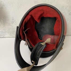 May include: A vintage-style open-face helmet featuring a smooth red outer shell and a contrasting black trim along the edge. The interior is lined with a vibrant red fabric padding, complemented by a black chin strap with a metal buckle mechanism.