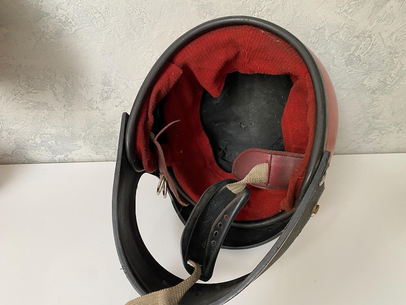 May include: A vintage-style open-face helmet featuring a smooth red outer shell and a contrasting black trim along the edge. The interior is lined with a vibrant red fabric padding, complemented by a black chin strap with a metal buckle mechanism.