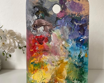 Op de afbeelding: Een houten kunstenaarspalet, rechthoekig met een duimgat, bedekt met een levendige reeks olieverf. De kleuren zijn rood, geel, blauw, groen, paars en wit, waardoor een gestructureerd, abstract ontwerp ontstaat.
